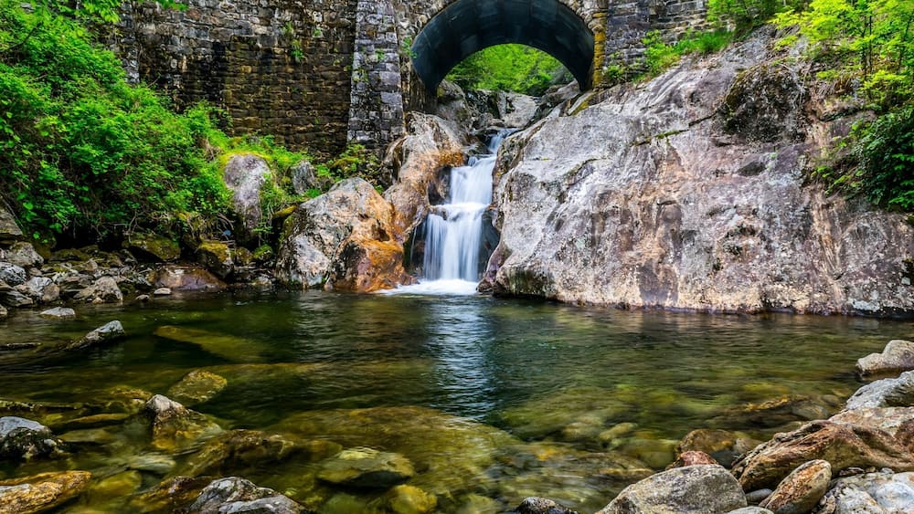 Un pont de pierre enjambe un ruisseau aux eaux claires, avec une cascade, le tout étant entouré d’une végétation luxuriante.