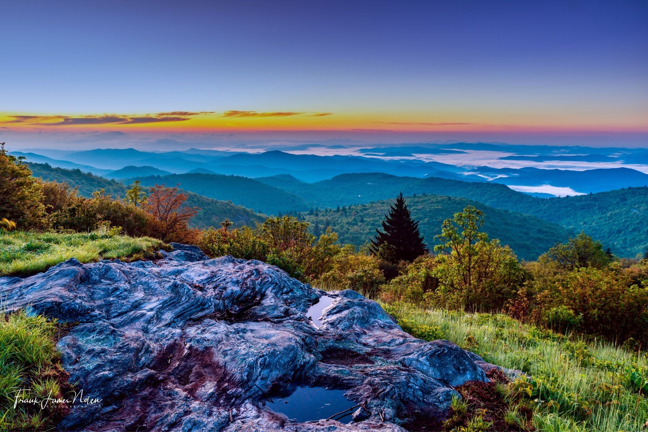A mountainous landscape with a large rock in the foreground and a view of distant hills under a colorful sunset sky.