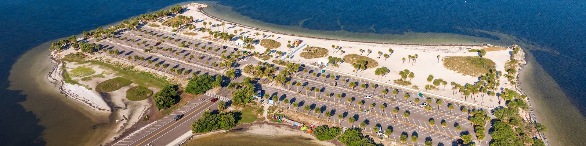 Aerial view of Fred Howard Park in Tarpon Springs, Florida, featuring parking lots, palm trees, sandy beaches, and surrounding blue waters under a clear sky.