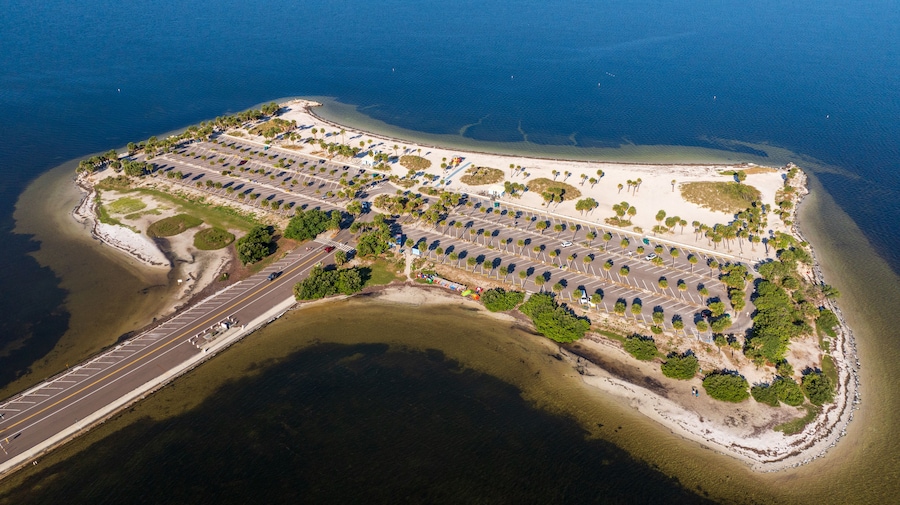 Aerial view of Fred Howard Park in Tarpon Springs, Florida, featuring parking lots, palm trees, sandy beaches, and surrounding blue waters under a clear sky.