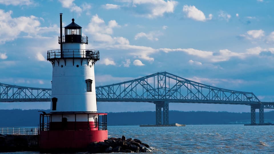 A lighthouse with a red and white structure, a metal bridge in the background, and calm water.