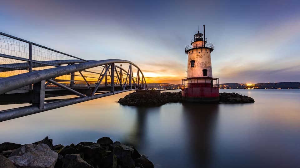 A lighthouse, a metal bridge, and calm water at sunset.