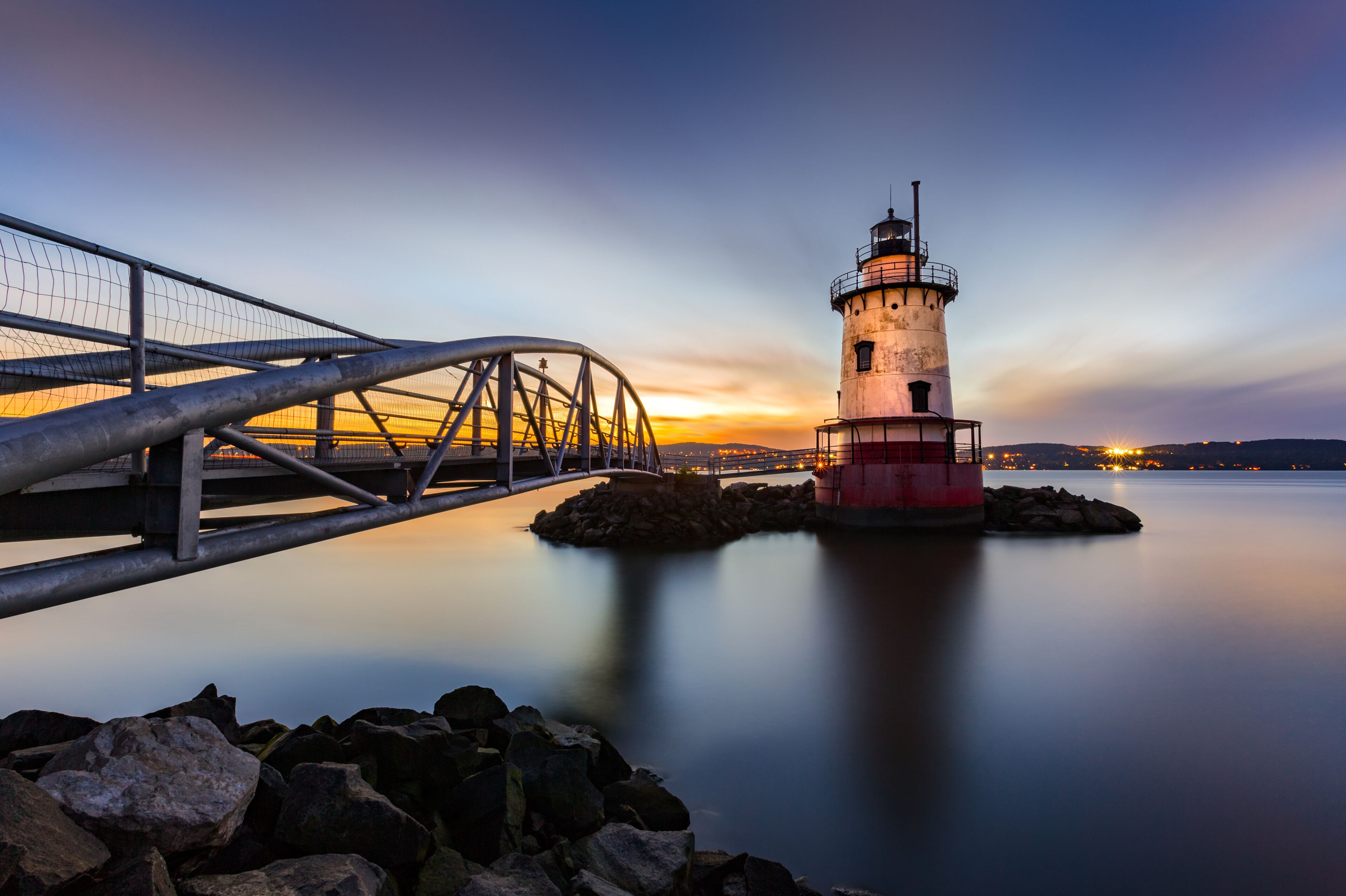 A lighthouse, a metal bridge, and calm water at sunset.