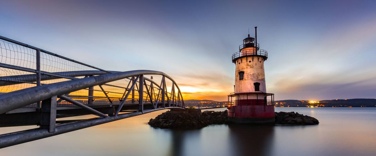 A lighthouse, a metal bridge, and calm water at sunset.