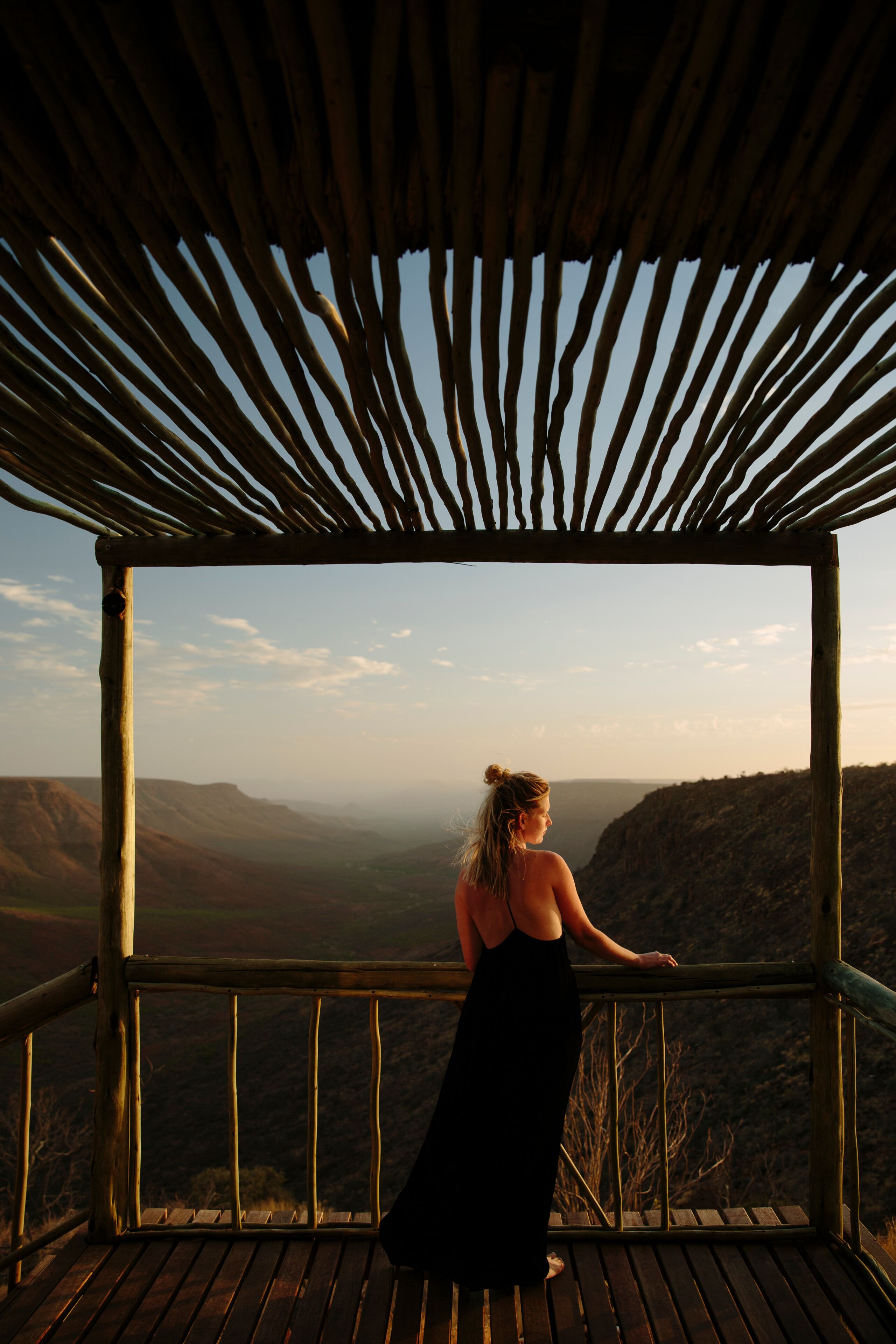 A person in a black dress standing on a wooden deck with a view of a mountainous landscape.
