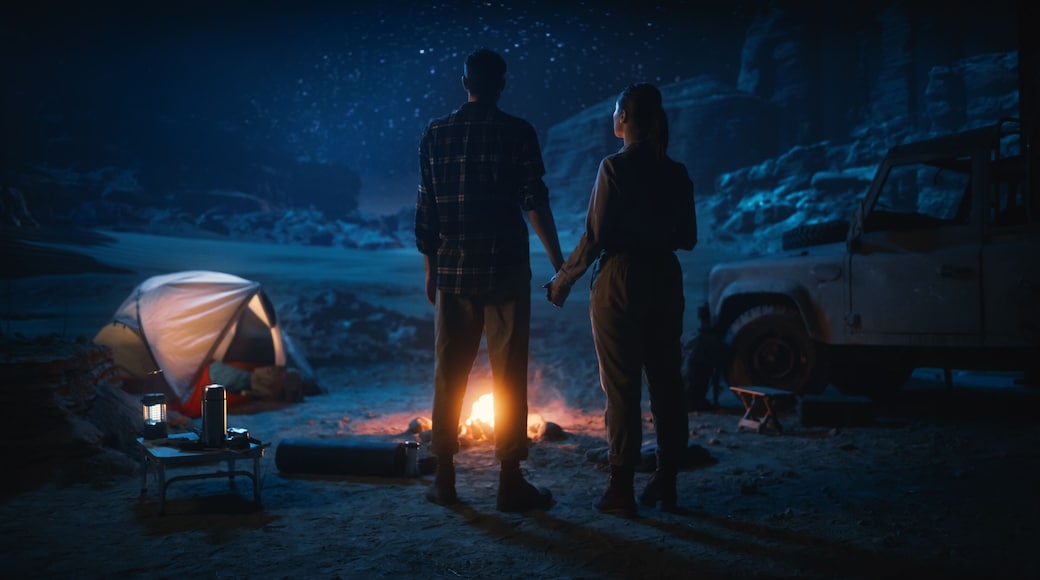 A couple standing by a campfire in a snowy landscape with a tent and a vehicle nearby.