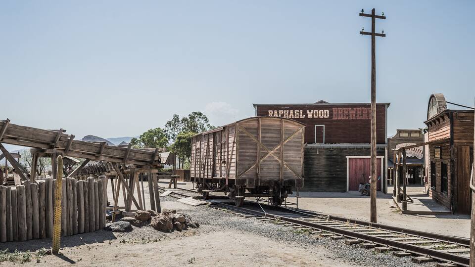 A vintage train station with a wooden train car labeled "Raphael Wood Brewery" and a cactus in the foreground.