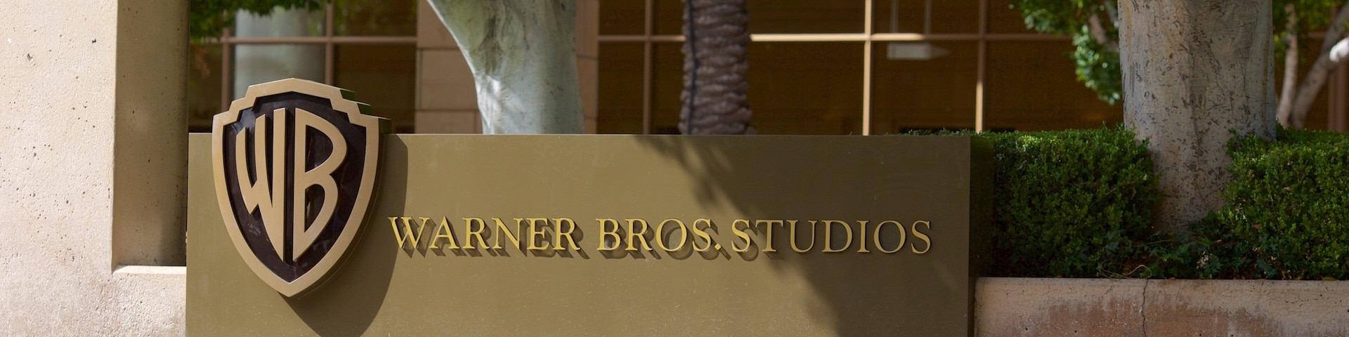 Placa da Warner Bros. Studios com logotipo, cercada por árvores e área ajardinada.