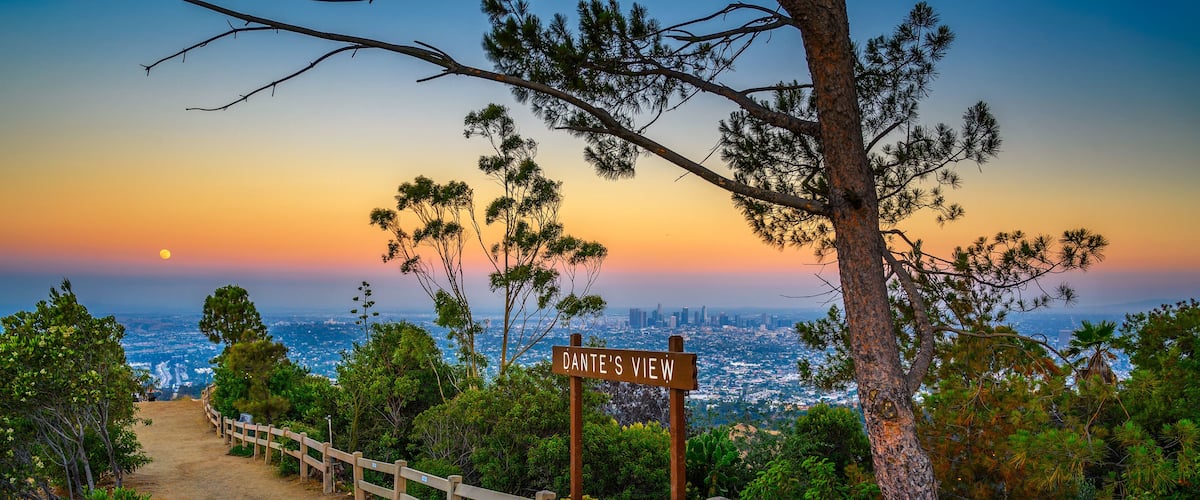 Un sendero de tierra con una valla de madera, un cartel que dice 'Dante's View' y un paisaje urbano al fondo durante el atardecer.
