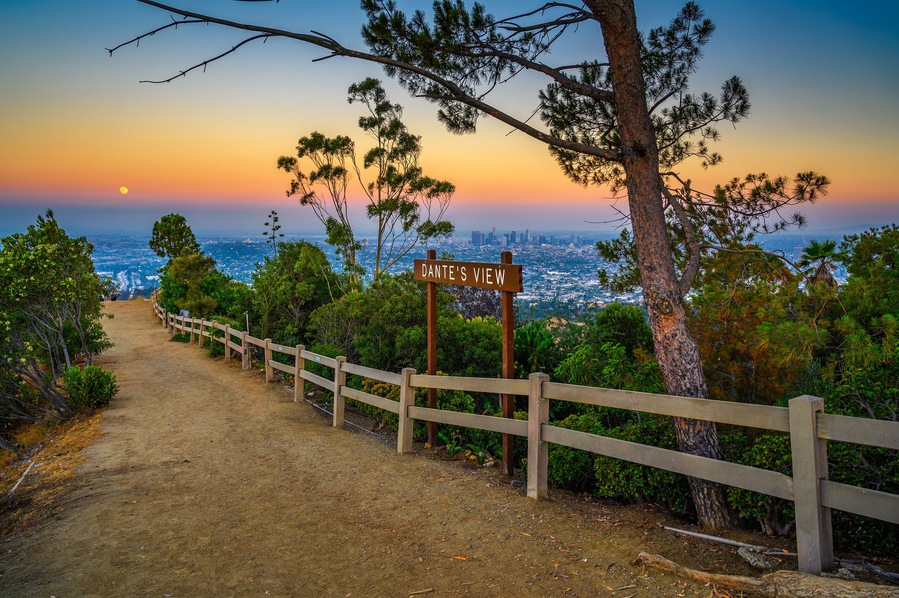 A dirt path with a wooden fence, a sign reading 'Dante's View,' and a cityscape in the background during sunset.