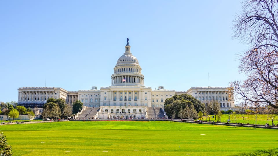 A large government building with a prominent dome and flag, surrounded by a well-maintained lawn and trees.