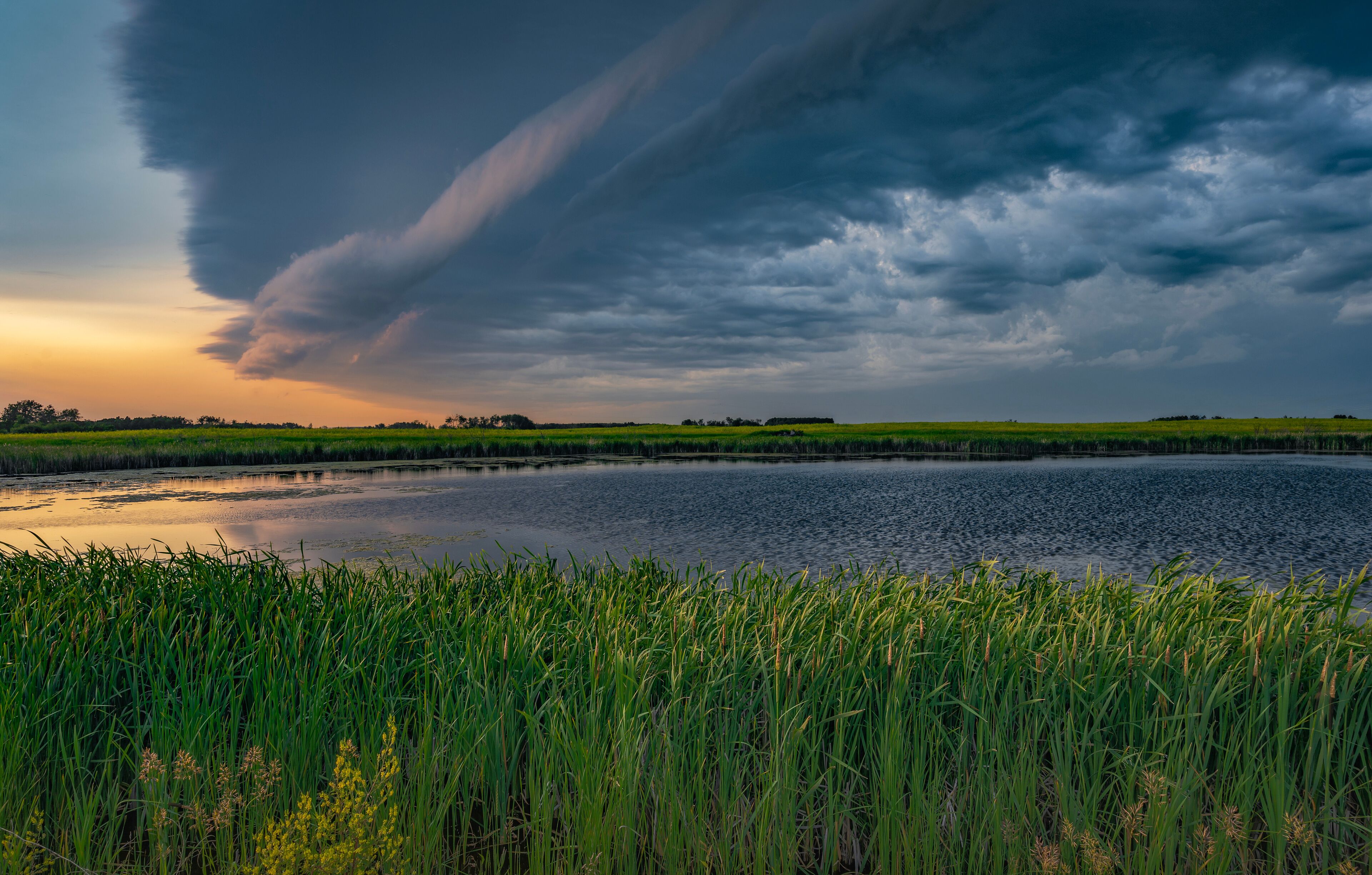 A stormy sky over a calm lake with green fields in the foreground.
