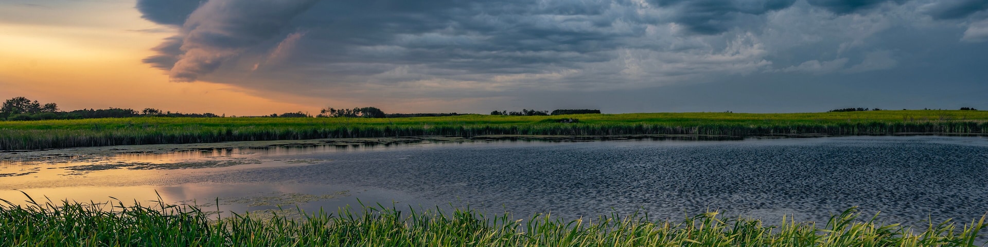 A stormy sky over a calm lake with green fields in the foreground.