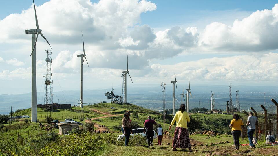 A landscape with wind turbines, a clear sky, and people walking on a grassy hill.