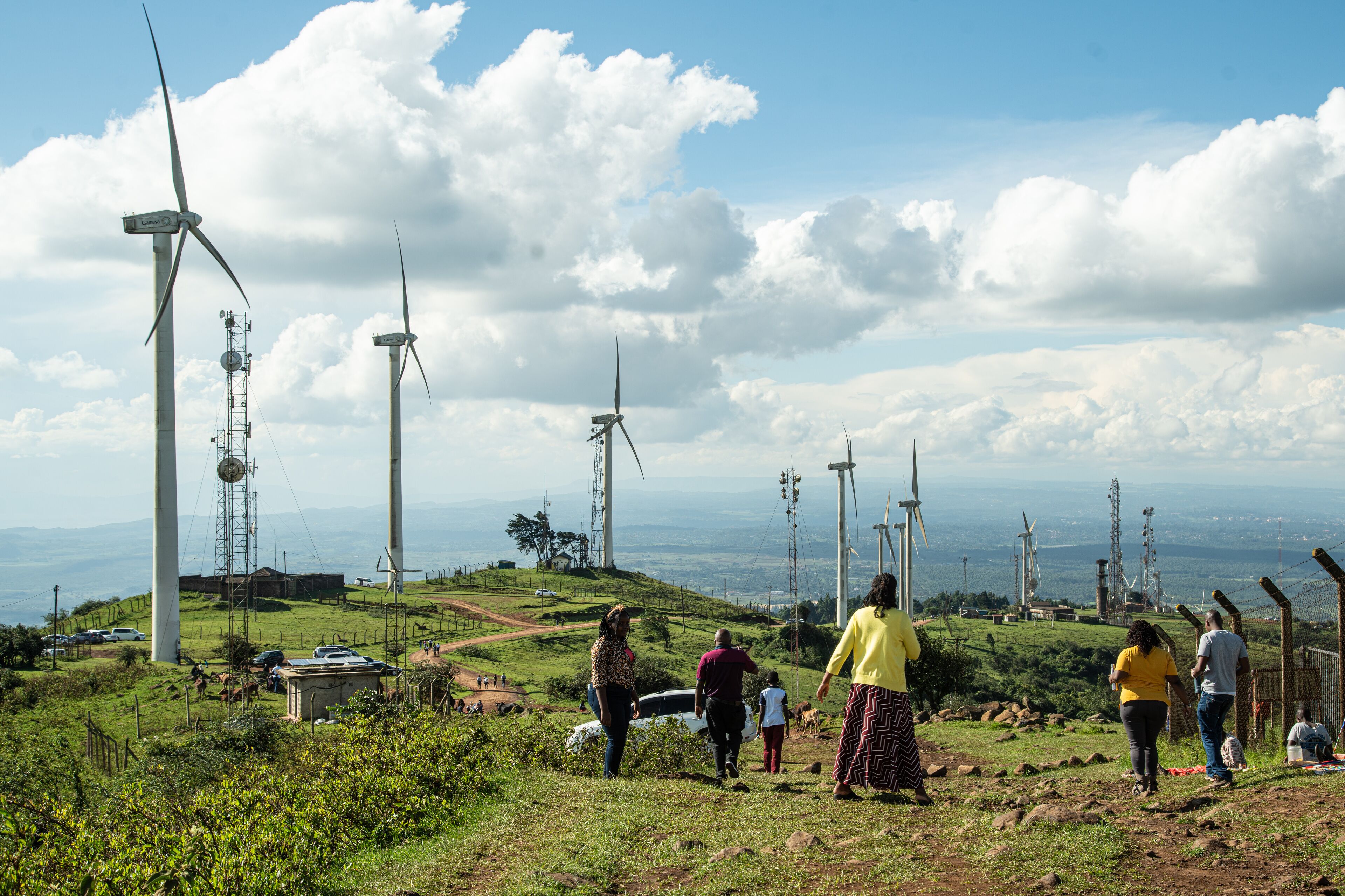 A landscape with wind turbines, a clear sky, and people walking on a grassy hill.