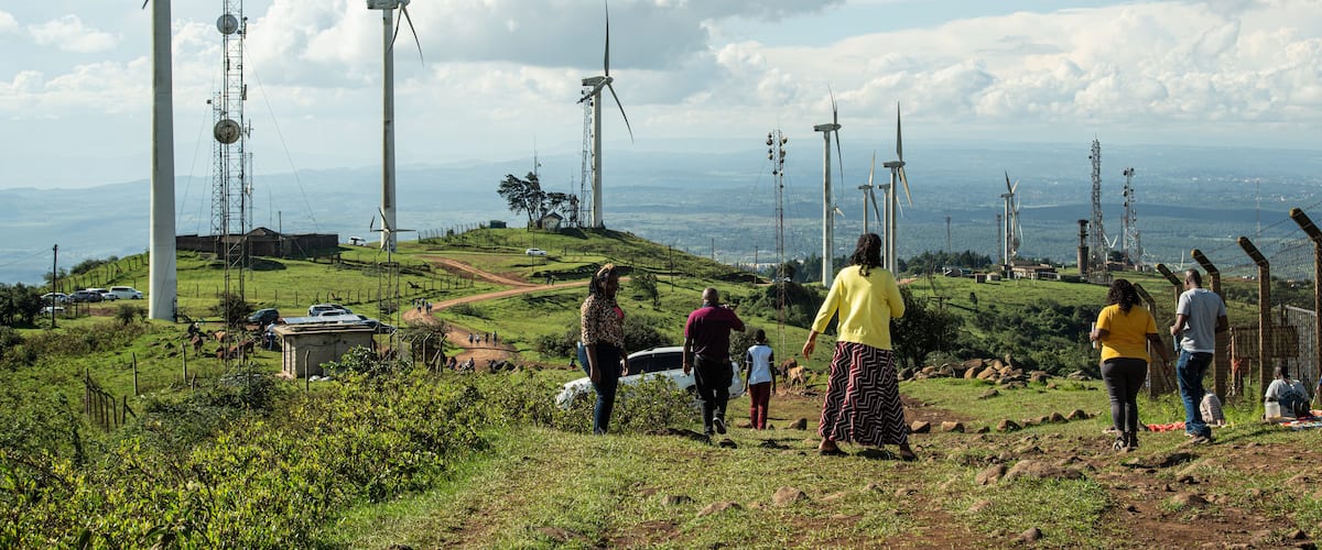 A landscape with wind turbines, a clear sky, and people walking on a grassy hill.