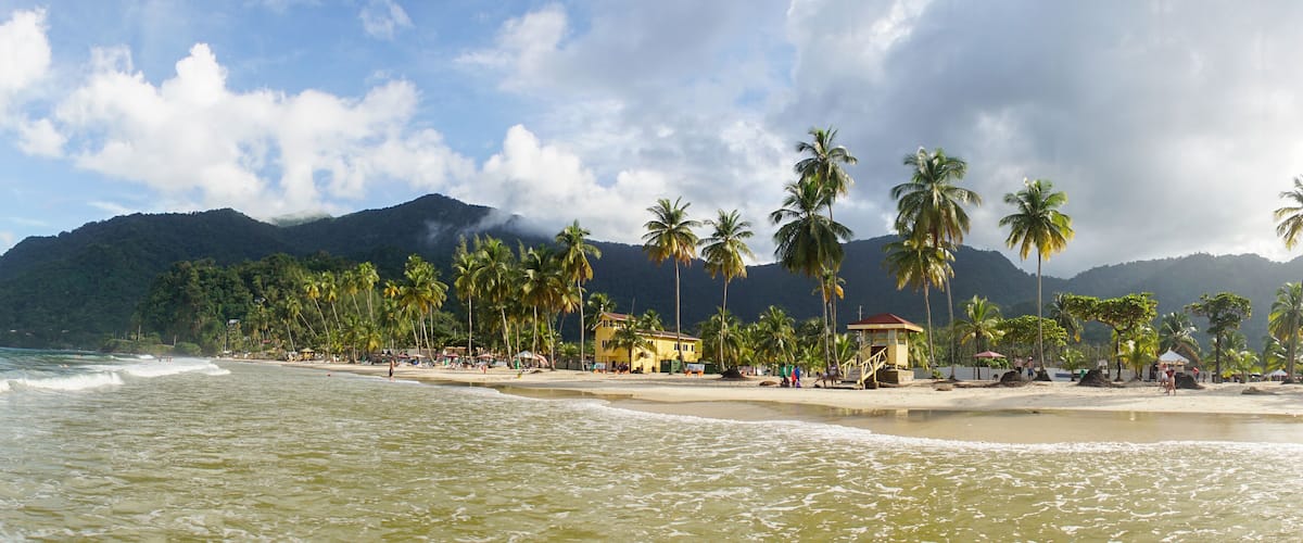 A beach with palm trees, a yellow building, and mountains in the background.