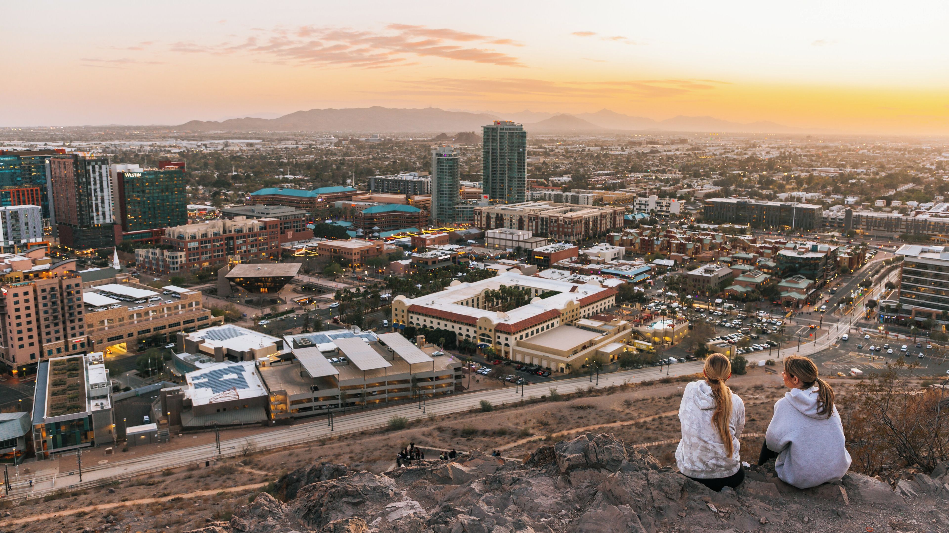 A cityscape at sunset with prominent buildings, a clear sky, and a distant mountain range.