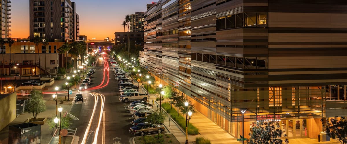 A city street at dusk with illuminated buildings and parked cars.