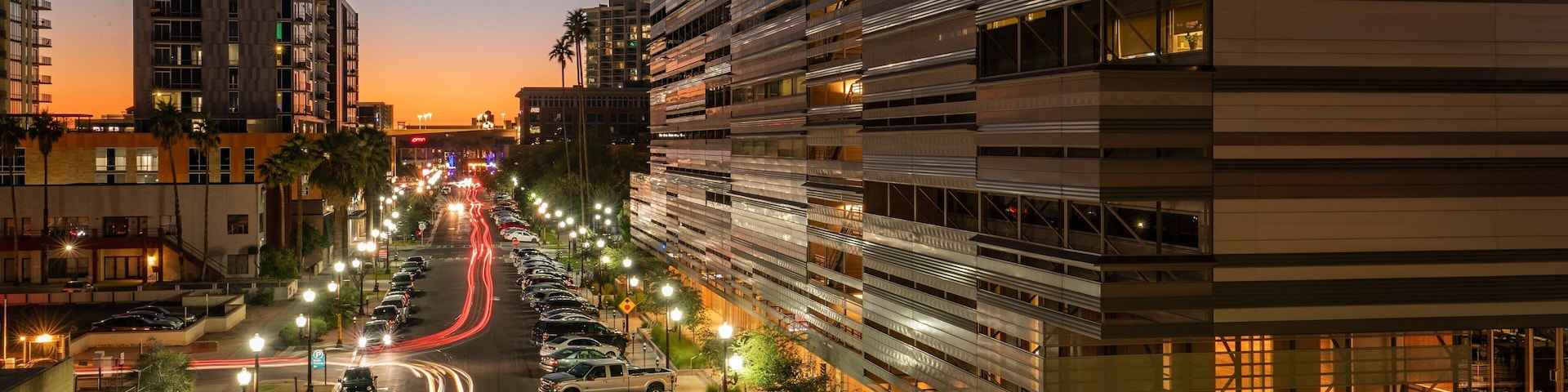 A city street at dusk with illuminated buildings and parked cars.