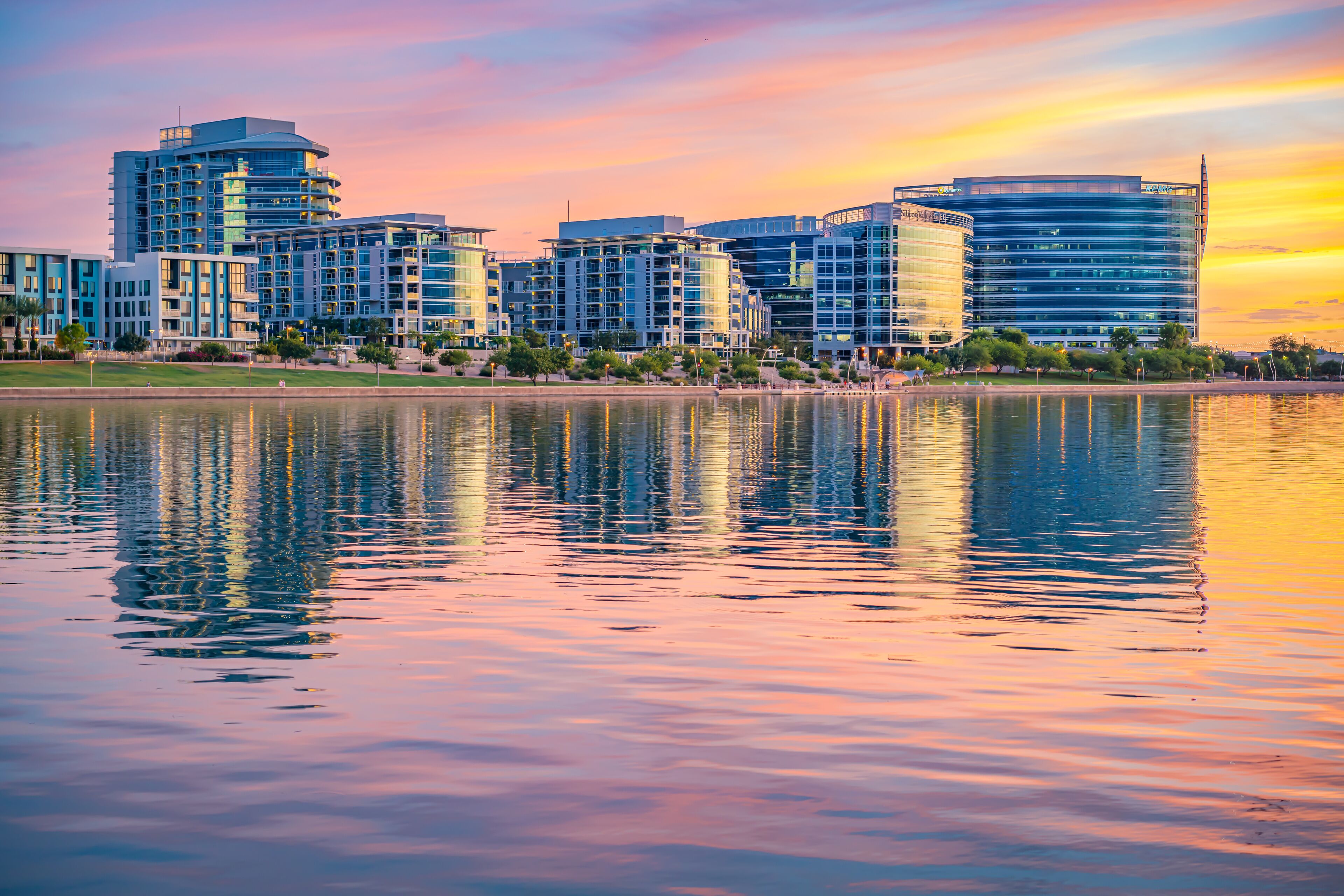 A cityscape with modern buildings and a calm lake reflecting the sunset.