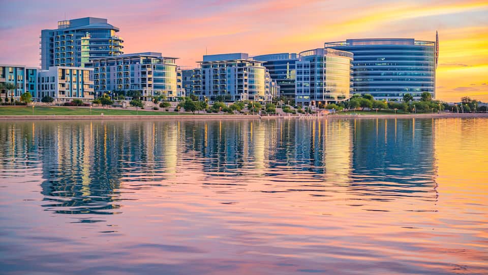 A cityscape with modern buildings and a calm lake reflecting the sunset.