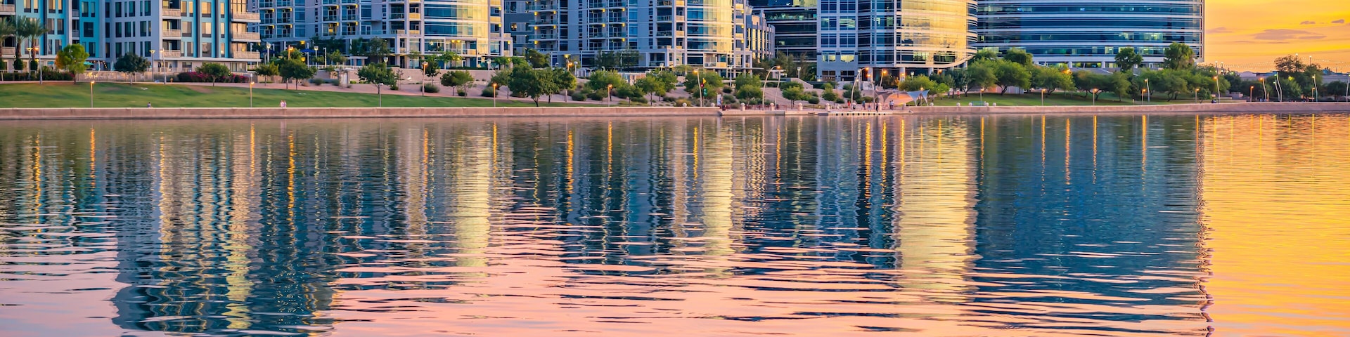 Paisagem urbana com edifícios modernos e um lago tranquilo refletindo o pôr do sol.