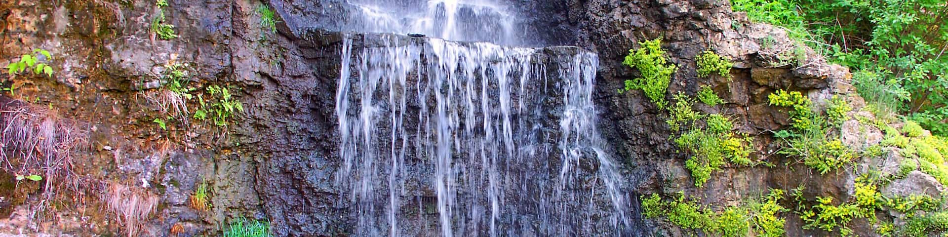 A multi-tiered waterfall cascading down a rocky cliff surrounded by lush greenery.