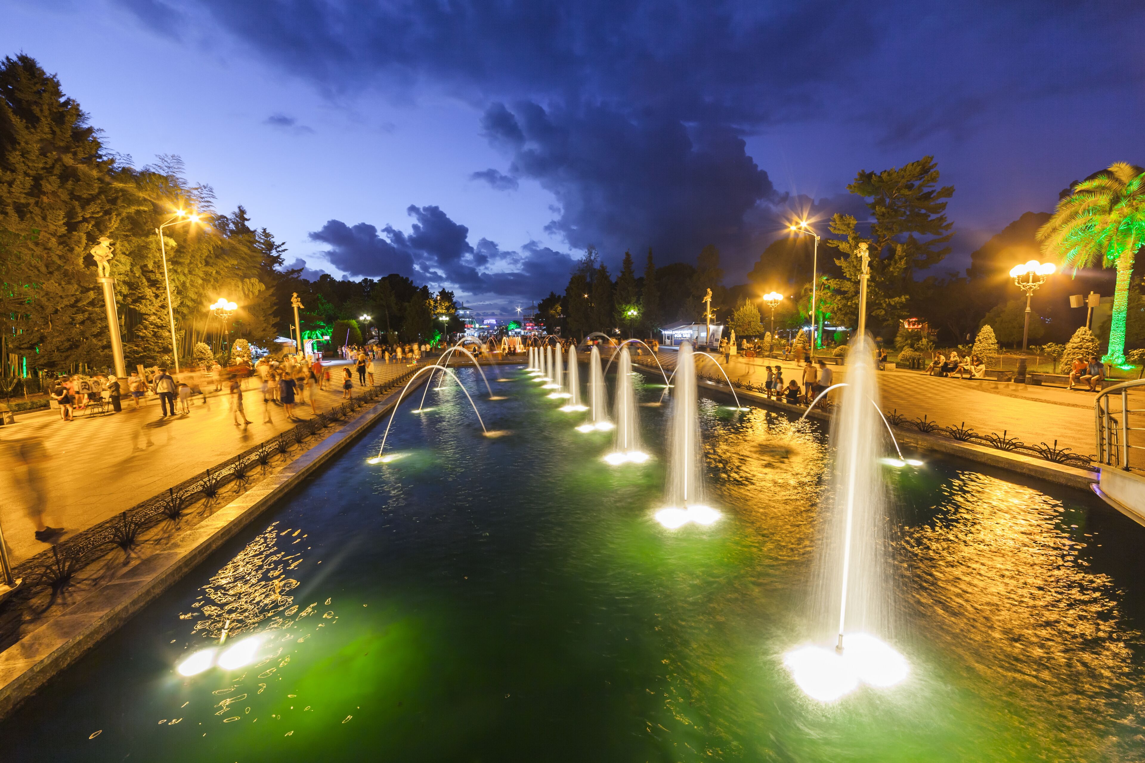 Un espectáculo nocturno de fuentes con múltiples chorros de agua, rodeado de un paseo bien iluminado y árboles.