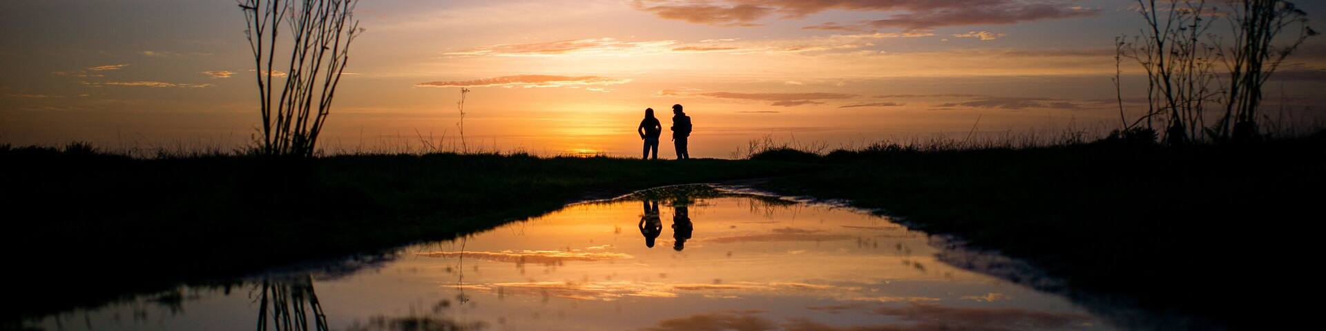 A sunset over a reflective water body with silhouettes of two people and sparse vegetation.