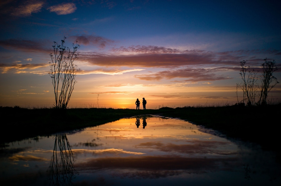Pôr do sol refletido na água, com silhuetas de duas pessoas e vegetação rasteira.