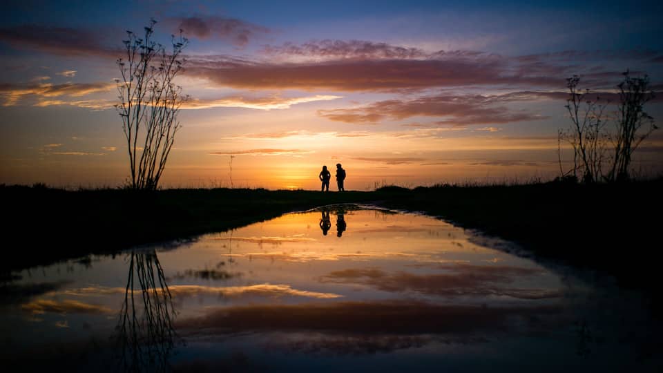 A sunset over a reflective water body with silhouettes of two people and sparse vegetation.