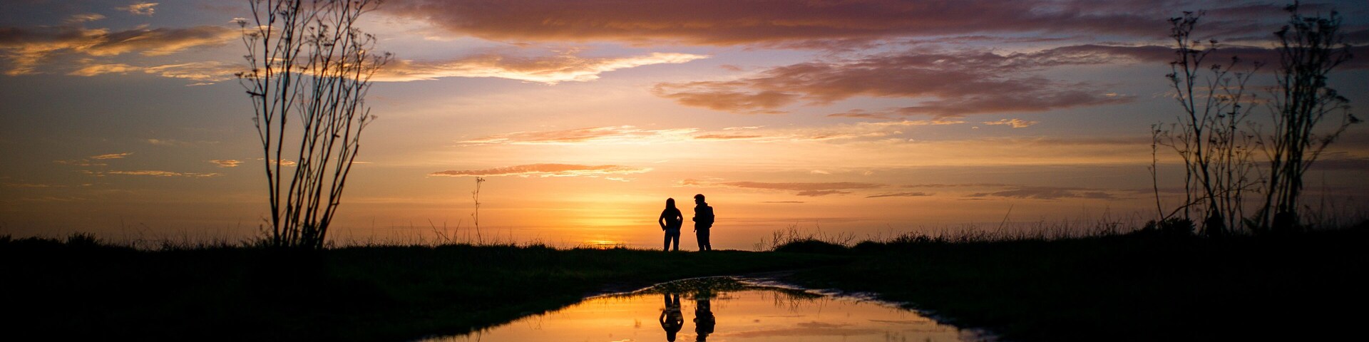 Pôr do sol refletido na água, com silhuetas de duas pessoas e vegetação rasteira.