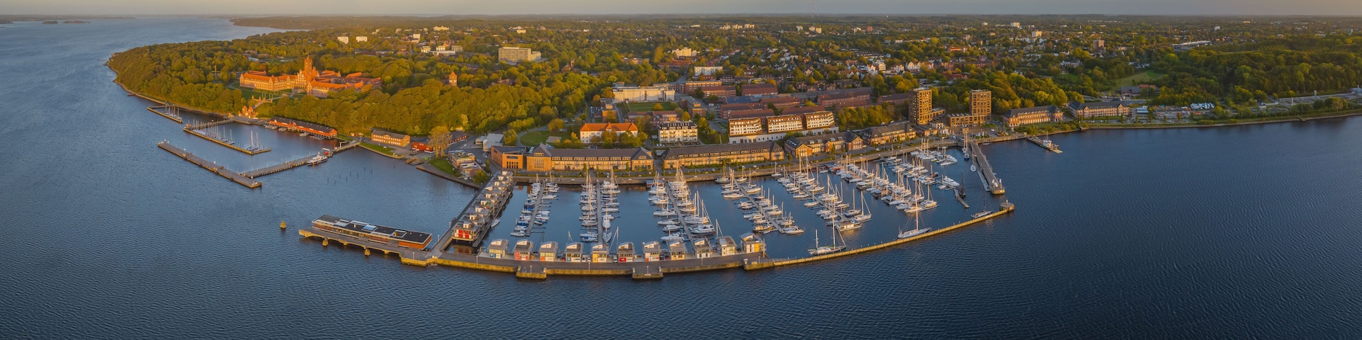 An aerial view of a marina with boats docked, surrounded by a cityscape and a forested island.