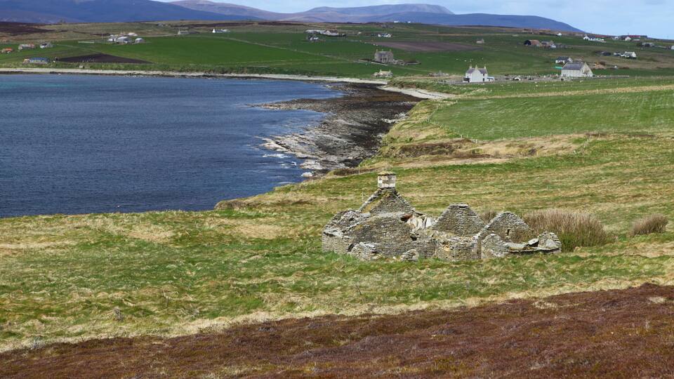 A coastal landscape with a stone ruin, grassy fields, and scattered houses.