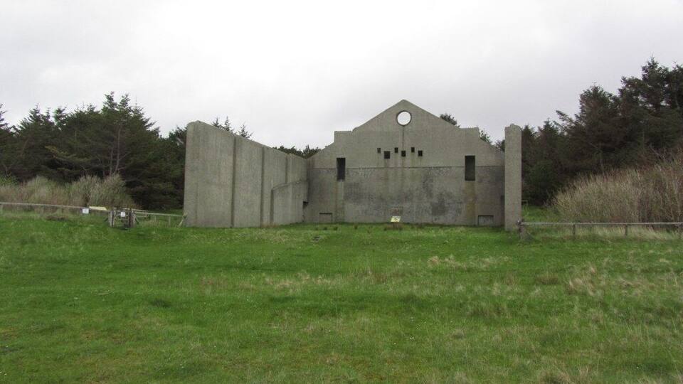 A concrete structure with a triangular roof and circular window, surrounded by a grassy field and trees.