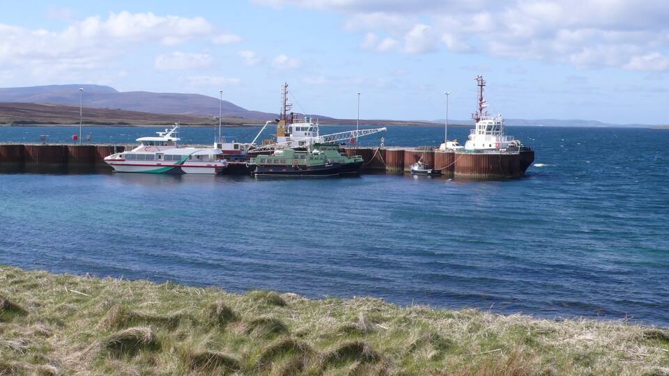 A harbor with three boats docked at a pier, surrounded by grassy hills and a clear sky.