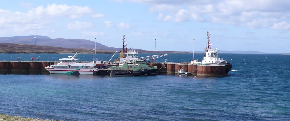 A harbor with three boats docked at a pier, surrounded by grassy hills and a clear sky.