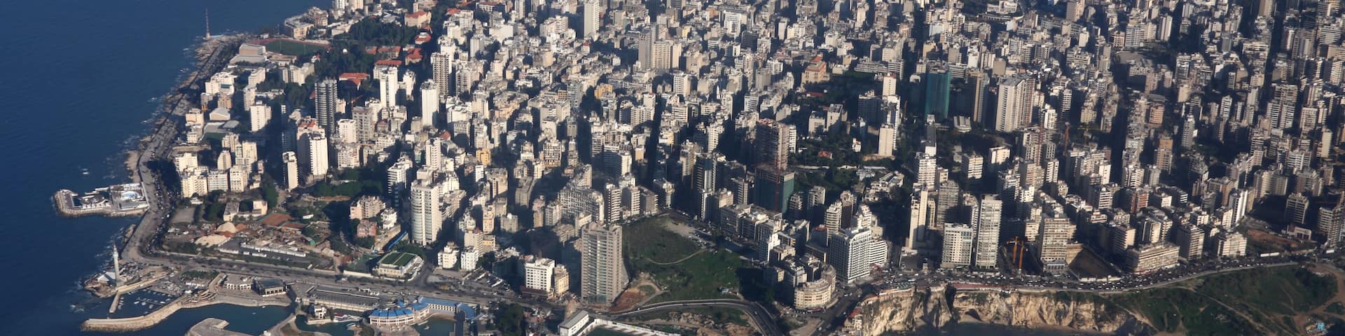 An aerial view of a densely populated coastal city with a prominent harbor, a large stadium, and a rocky coastline.