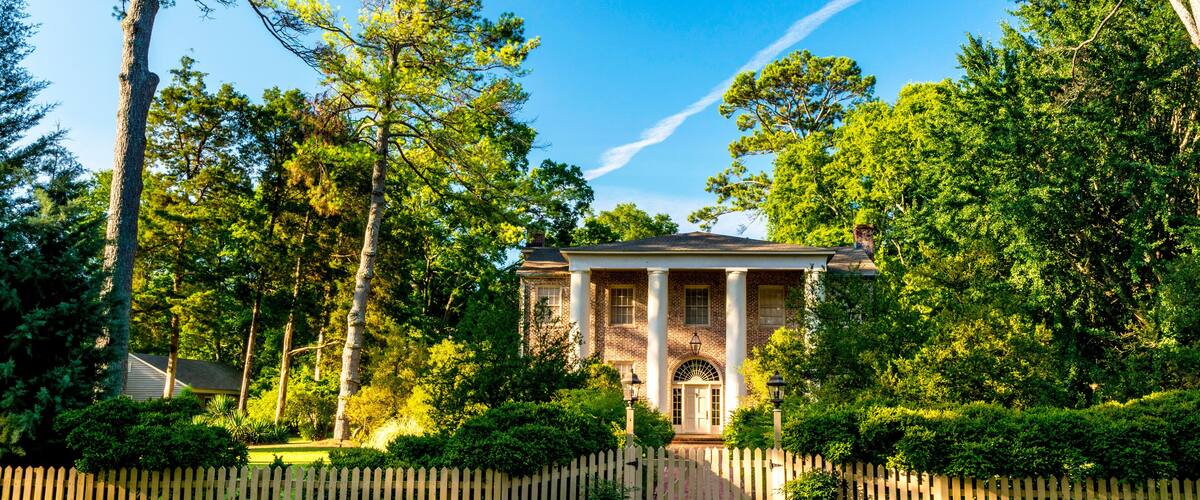 A historic building with columns, surrounded by lush greenery and a wooden fence.