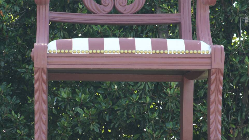 A maroon chair with a decorative backrest and a striped cushion, set against a backdrop of dense green foliage.