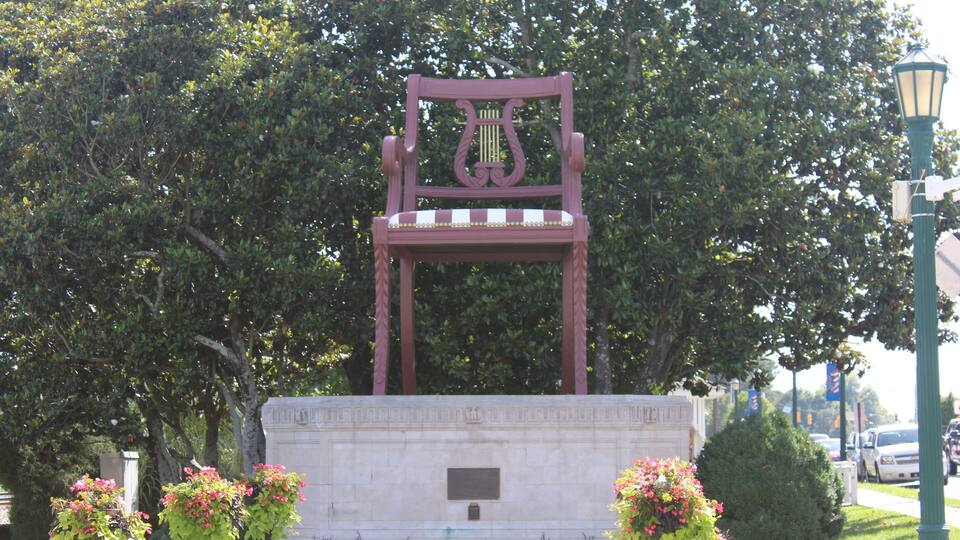 A large pink chair sculpture in a park with trees and flowers.