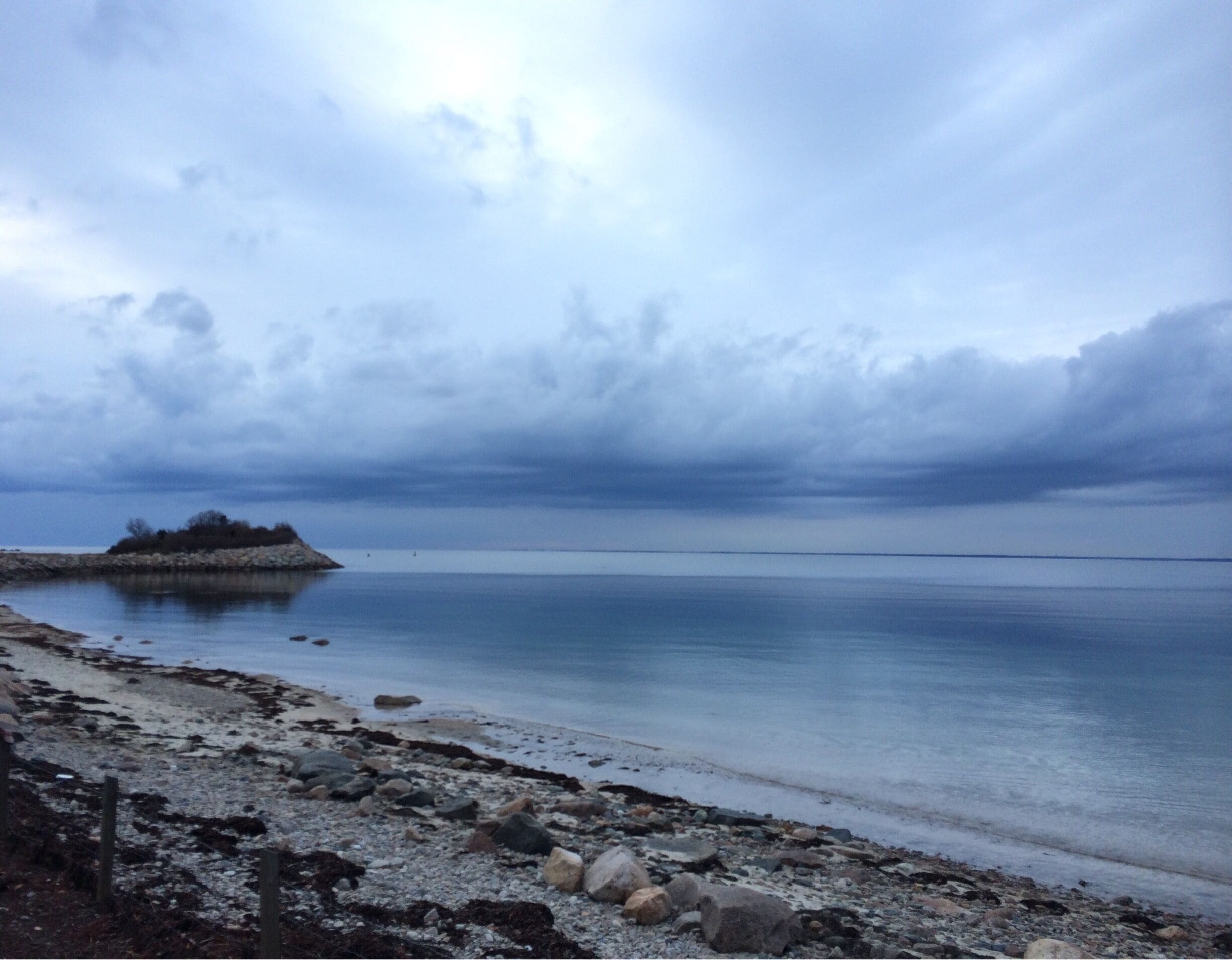 A rocky shoreline with calm water and a small island in the distance under a cloudy sky.