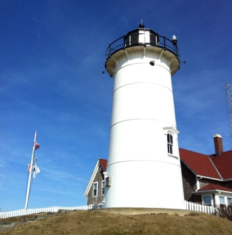 A white lighthouse with a black top, a white picket fence, and a red-roofed house in the background.