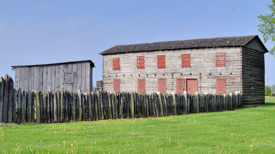 A rustic wooden building with red shutters and a wooden fence in a grassy field.