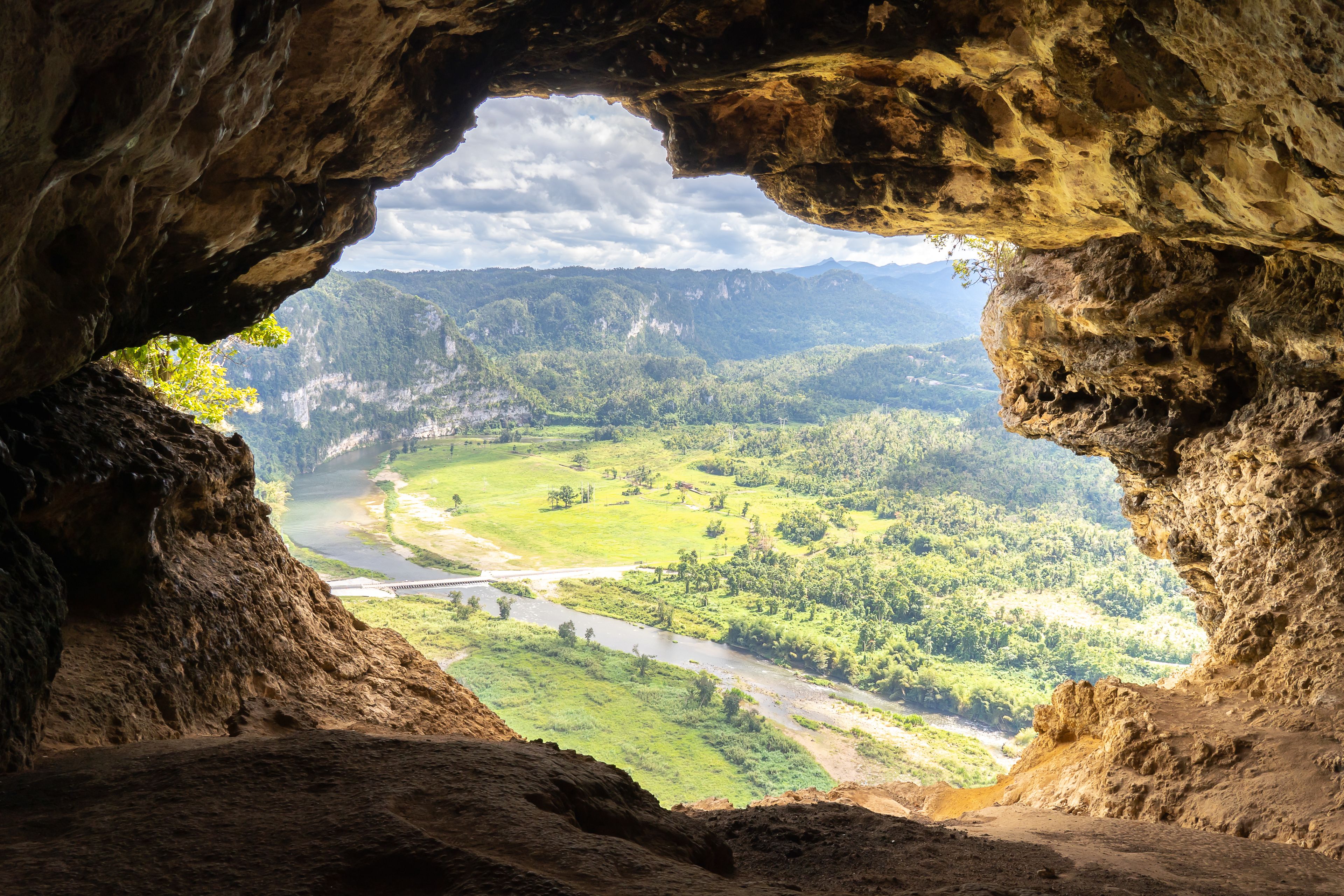 Boca de cueva con vista a un río, un valle verde y montañas distantes.