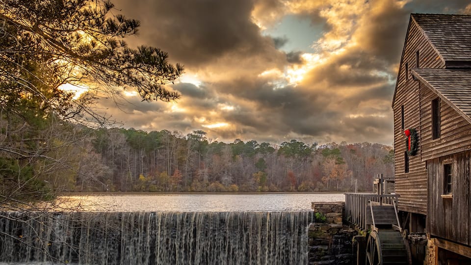 A wooden mill with a water wheel, a waterfall, and a forested shoreline.