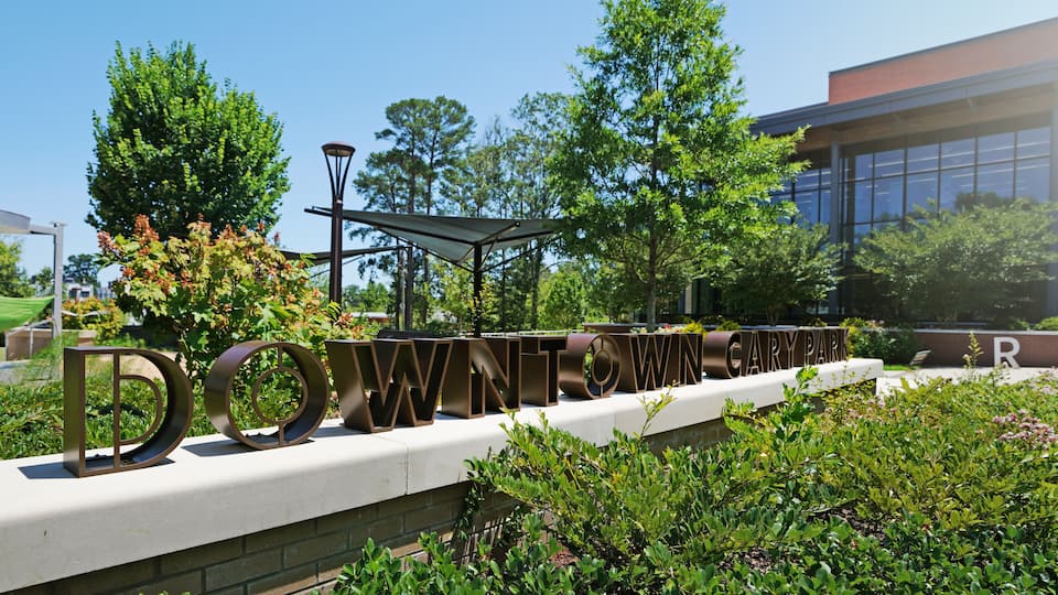 A sign with 'DOWNTOWN' in large letters, surrounded by greenery and a building with large windows.