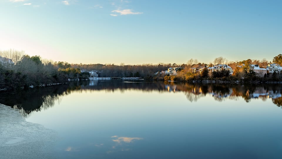 A calm lake with reflections of houses and trees, surrounded by a forested area.