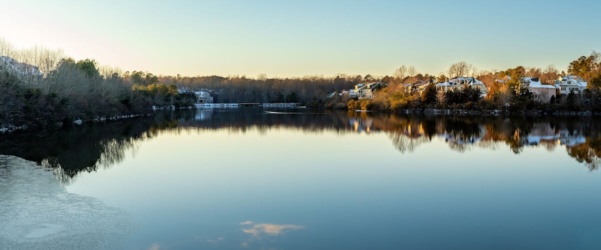 A calm lake with reflections of houses and trees, surrounded by a forested area.